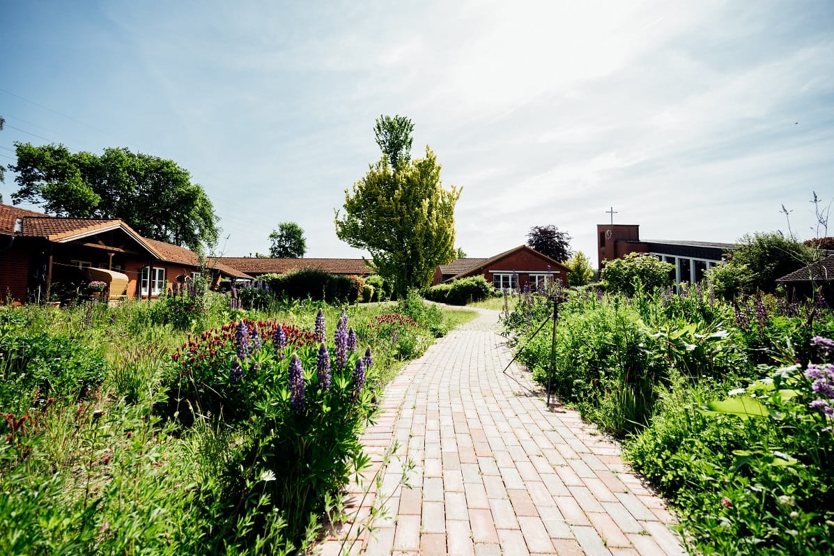 Ein gepflasterter Weg führt durch den blühenden Garten zu den Wohnhäusern.
