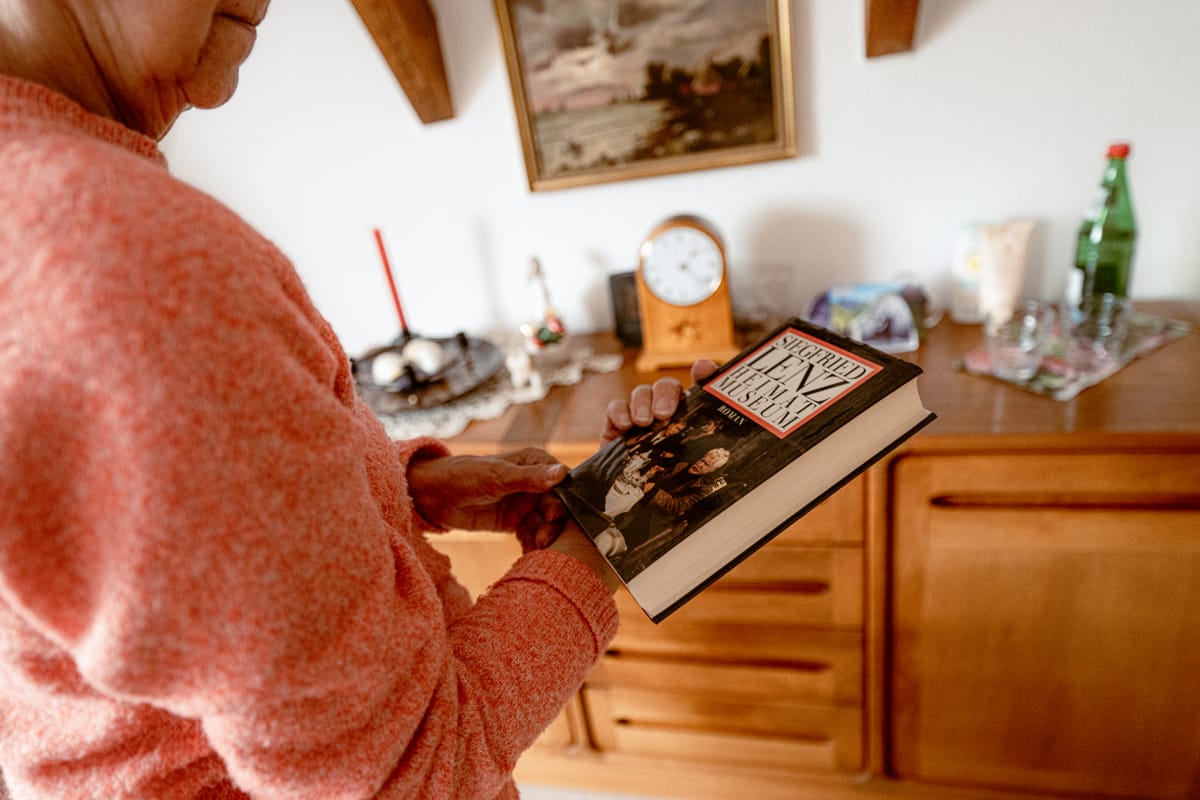 Eine Bewohnerin im roten Pullover blättert in einem Buch, dahinter eine Kommode mit Uhr.
